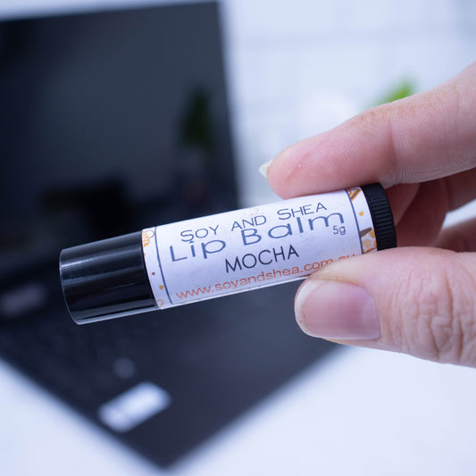 A hand holds a black tube of lip balm against a blurred background with an open laptop computer on a white surface.  The label has text LIP BALM MOCHA on a white background with a board of cups filled with coffee.