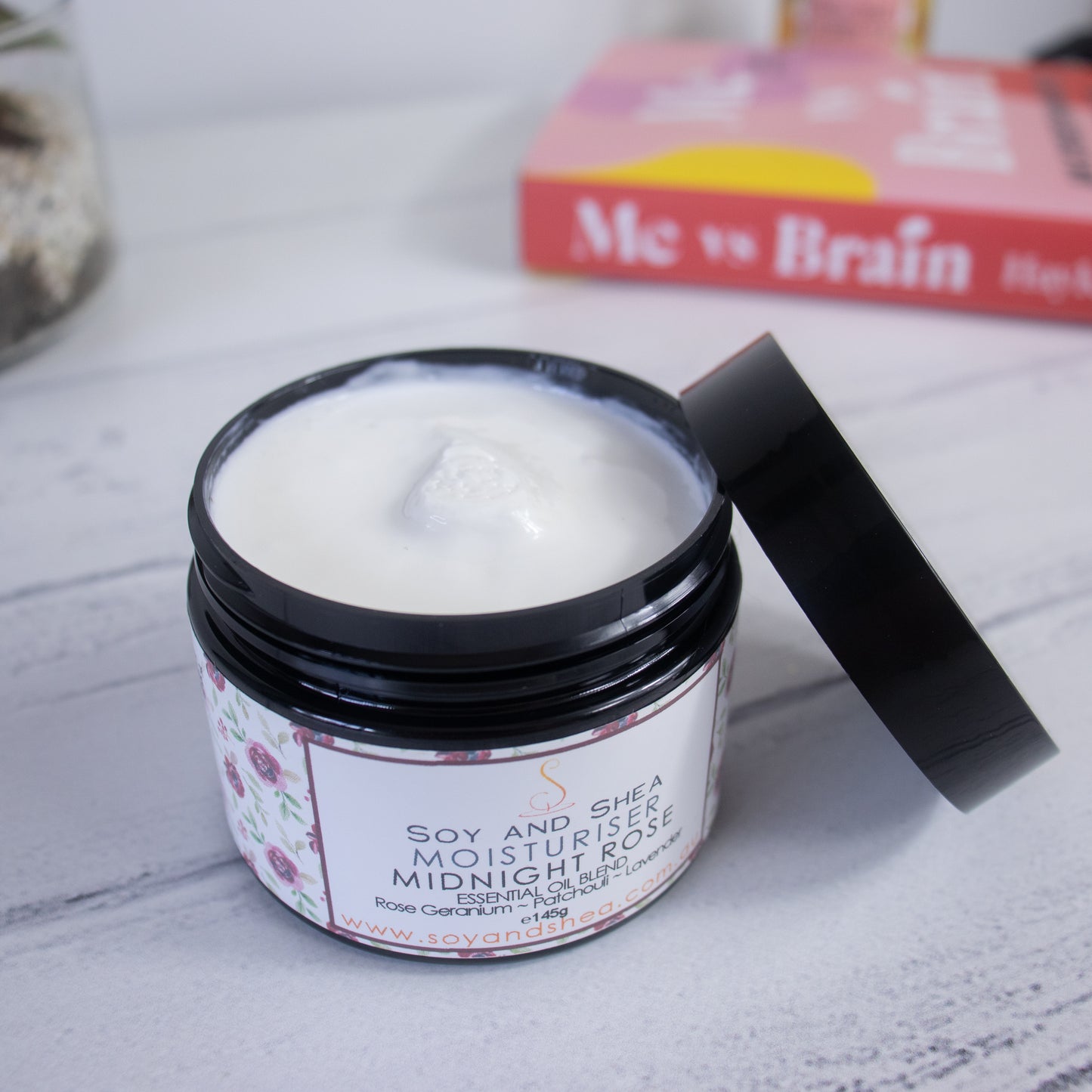 A black jar with an open lid containing white cream labeled "Midnight Rose Moisturiser (Essential Oil Blend)" by Soy and Shea sits on a white wooden surface. With notes of Patchouli, this body moisturizer promises a luxurious feel. In the blurred background, there's a book titled "Me vs Brain", featuring vibrant colors.