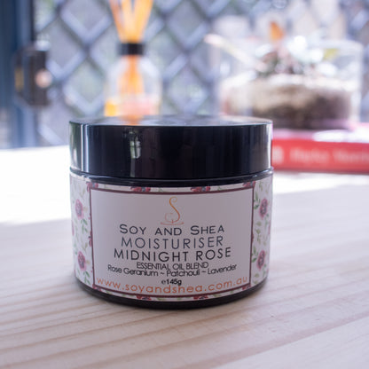 A jar of Soy and Shea's Midnight Rose Moisturiser (Essential Oil Blend) rests on a wooden surface. The label highlights a blend of essential oils, including rose geranium, patchouli, and lavender. In the blurred background, you can see some books and a potted plant. The jar contains 145 grams.