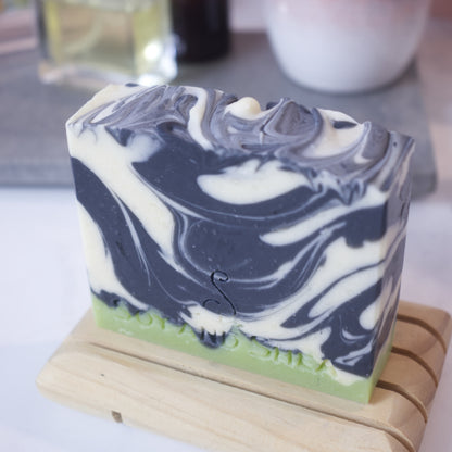  A bar of soap that has a small layer of green on the base with a black and white swirl pattern above, sits on a wooden soap dish with a blurred background