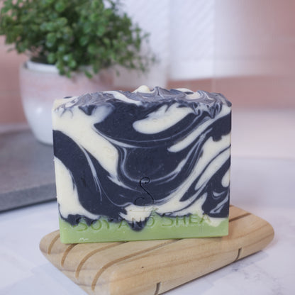 A bar of soap with a swirled pattern of black and white on a narrow layer of green is sitting on a wooden soap dish, with a potted plant and decorative items in the background.