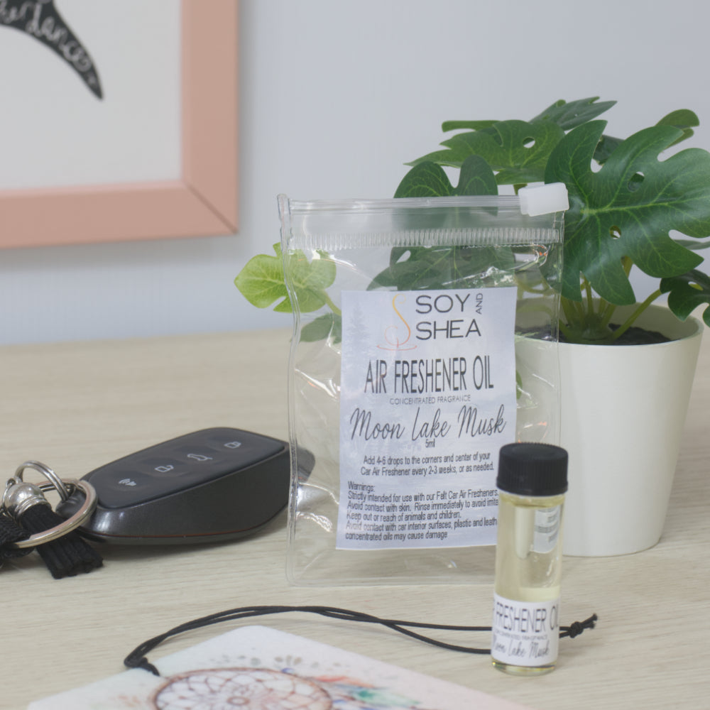 A small clear bottle with black cap of Soy's Shea Air Freshener Oil next to a clear resealable pouch, displayed on a light wood tabletop with a car key, a felt air freshener, and a potted plant in the background.  