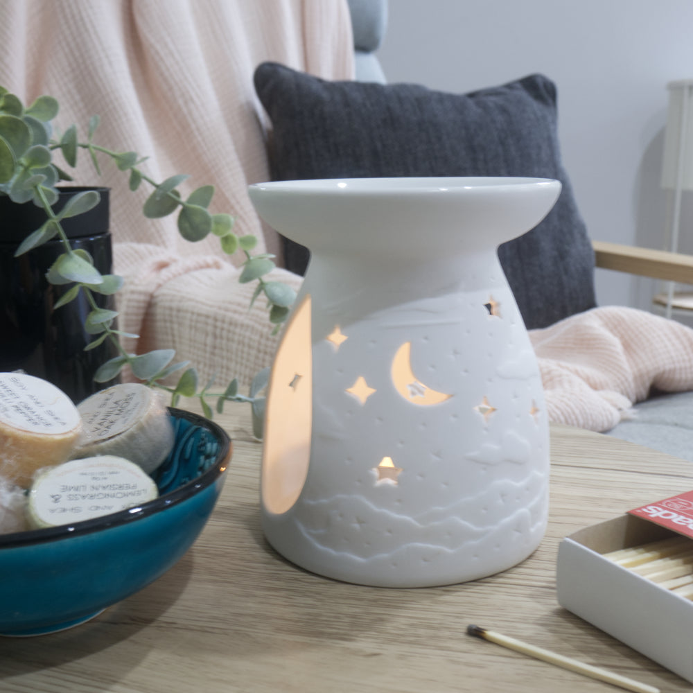 A matte white porcelain fragrance warmer sits on a light wooden table next to a blue bowl of wax melts and a box of matches. The warmer features embossed cloud details at the base and lit moon and stars cutouts that glow warmly from an internal tealight candle. In the blurred background, a pink textured throw blanket and a dark grey cushion rest on a chair