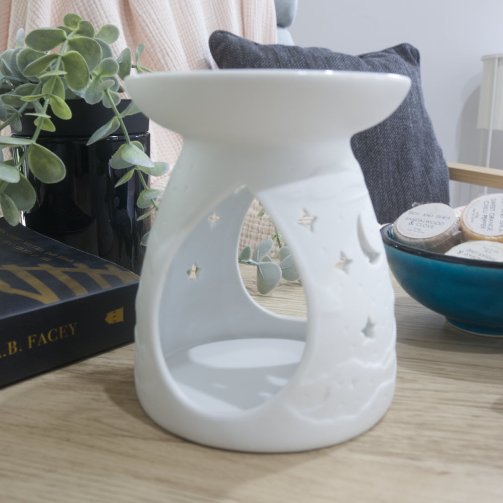 A matte white porcelain fragrance warmer sits on a light wooden table next to a blue bowl of wax melts and a box of matches. The image shows the warmer from the side showing large teardrop openings on the side of the warmer.