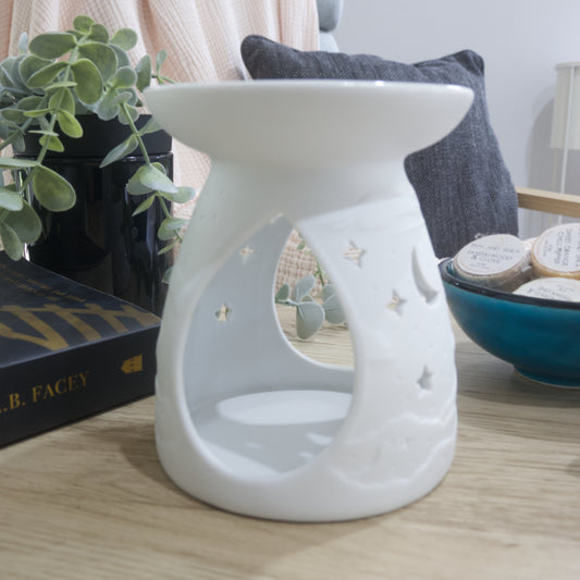 A matte white porcelain fragrance warmer sits on a light wooden table next to a blue bowl of wax melts and a box of matches. The image shows the warmer from the side showing large teardrop openings on the side of the warmer.