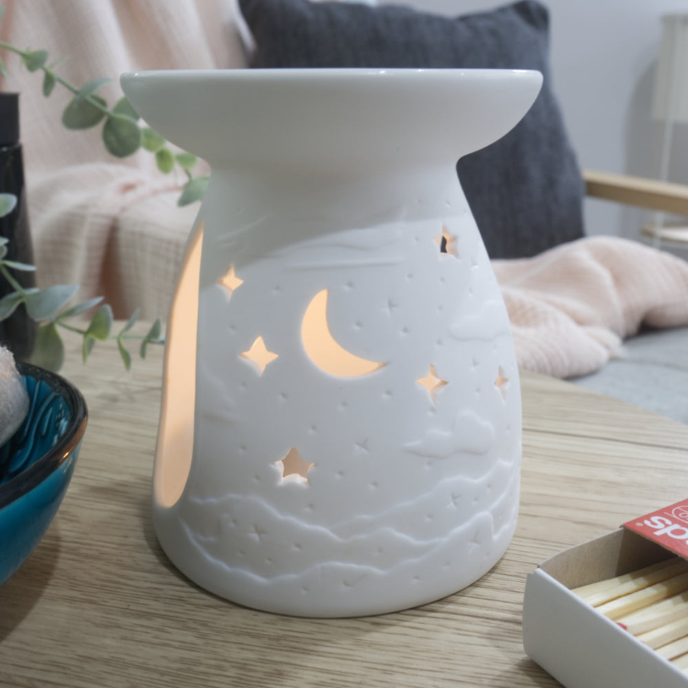 A matte white porcelain fragrance warmer sits on a light wooden table next to a blue bowl of wax melts and a box of matches. The warmer features embossed cloud details at the base and lit moon and stars cutouts that glow warmly from an internal tealight candle. In the blurred background, a pink textured throw blanket and a dark grey cushion rest on a chair