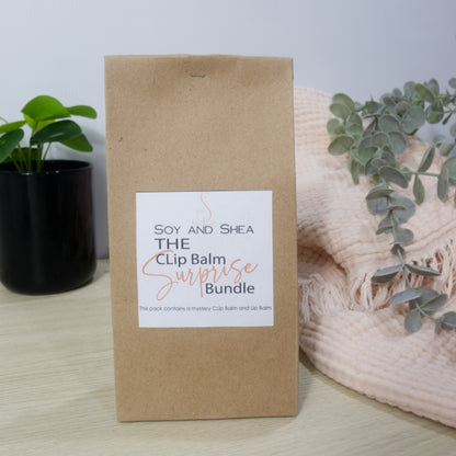 Kraft paper packaging with a label for 'The Clip Balm Surprise Bundle' by Soy and Shea, placed on a surface with a plant and blanket in the background.