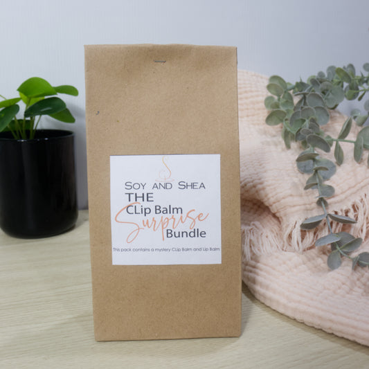 Kraft paper packaging with a label for 'The Clip Balm Surprise Bundle' by Soy and Shea, placed on a surface with a plant and blanket in the background.