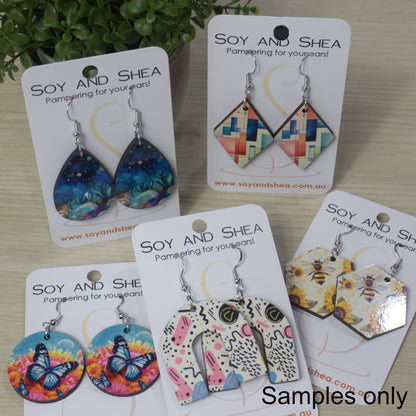 A collection of various earrings are displayed on their cardboard backing on a bench top.  The earrings are of various shapes and sizes with bright patterns.  All have silver hooks