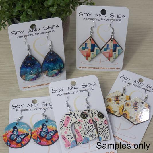 A collection of various earrings are displayed on their cardboard backing on a bench top.  The earrings are of various shapes and sizes with bright patterns.  All have silver hooks