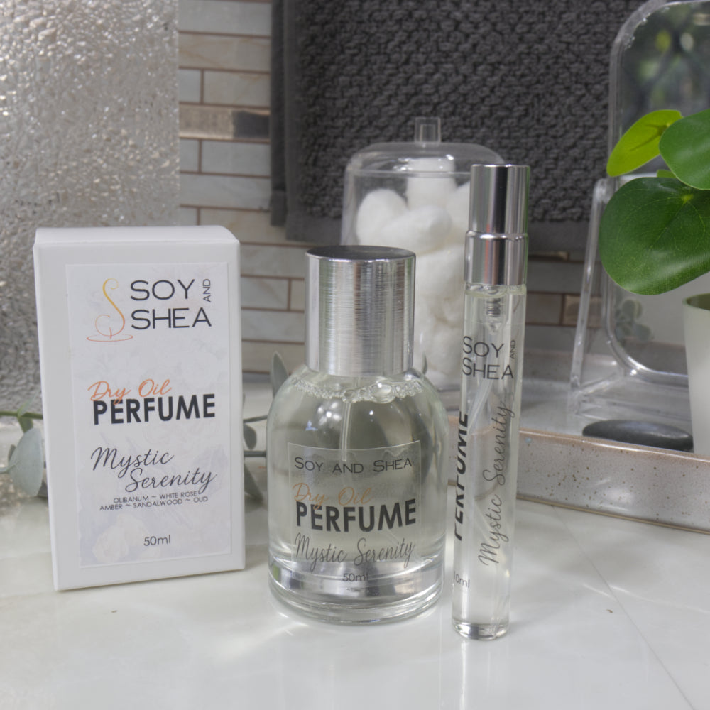 A selection of perfume bottles are presented on a bathroom countertop.  A clear, narrow, tall bottle labeled as 10ml with a silver cap stands beside a clear round, short bottle with silver lid labelled as 50ml.  Behind sits a white box with label identifying as Dry Oil Perfume Mystic Serenity.