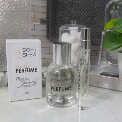 A selection of perfume bottles are presented on a bathroom countertop.  A clear, narrow, tall bottle labeled as 10ml with a silver cap stands beside a clear round, short bottle with silver lid labelled as 50ml.  Behind sits a white box with label identifying as Dry Oil Perfume Mystic Serenity.