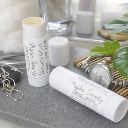 Two white tubes of Solid Perfume are displayed on a grey stone tray along with silver earrings, watch and greenery.   One tube lays flat  displaying the fragrance notes while another tube stands upright to the left with the lid removed and placed to the back revealing a beige balm