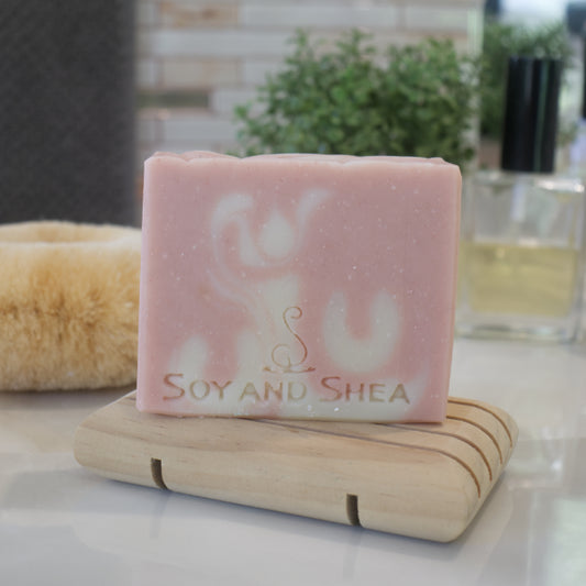 A bar of soap with a soft pink hue, featuring a white swirled design and placed on a wooden soap dish. The soap has the brand name 'Soy and Shea' inscribed on it.  The background is a blurred bathroom counter top