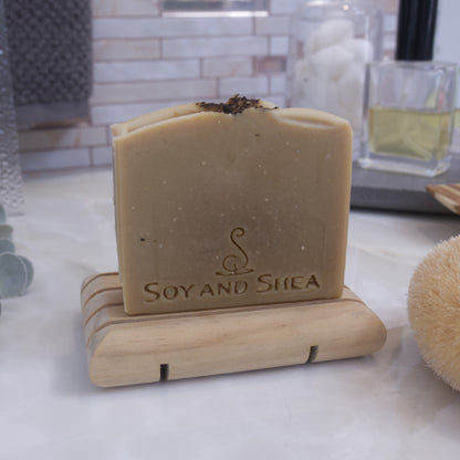 A pale khaki green sap bars stamped with SOY AND SHEA, sits on a wooden rack with a faded bathroom background.  Tea leaves are sprinkled on the top of the soap