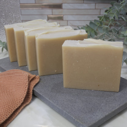 Five bars of creamy coloured soap sit in a row facing forward on a gray surface with a brown towel and greenery in the background.