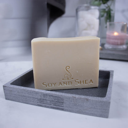 A cream bar of soap stamped with SOY AND SHEA stands on a grey stone soap dish on bathroom countertop.  In the background is a burning candle, jar of cotton balls and a towel hanging against a tile wall.