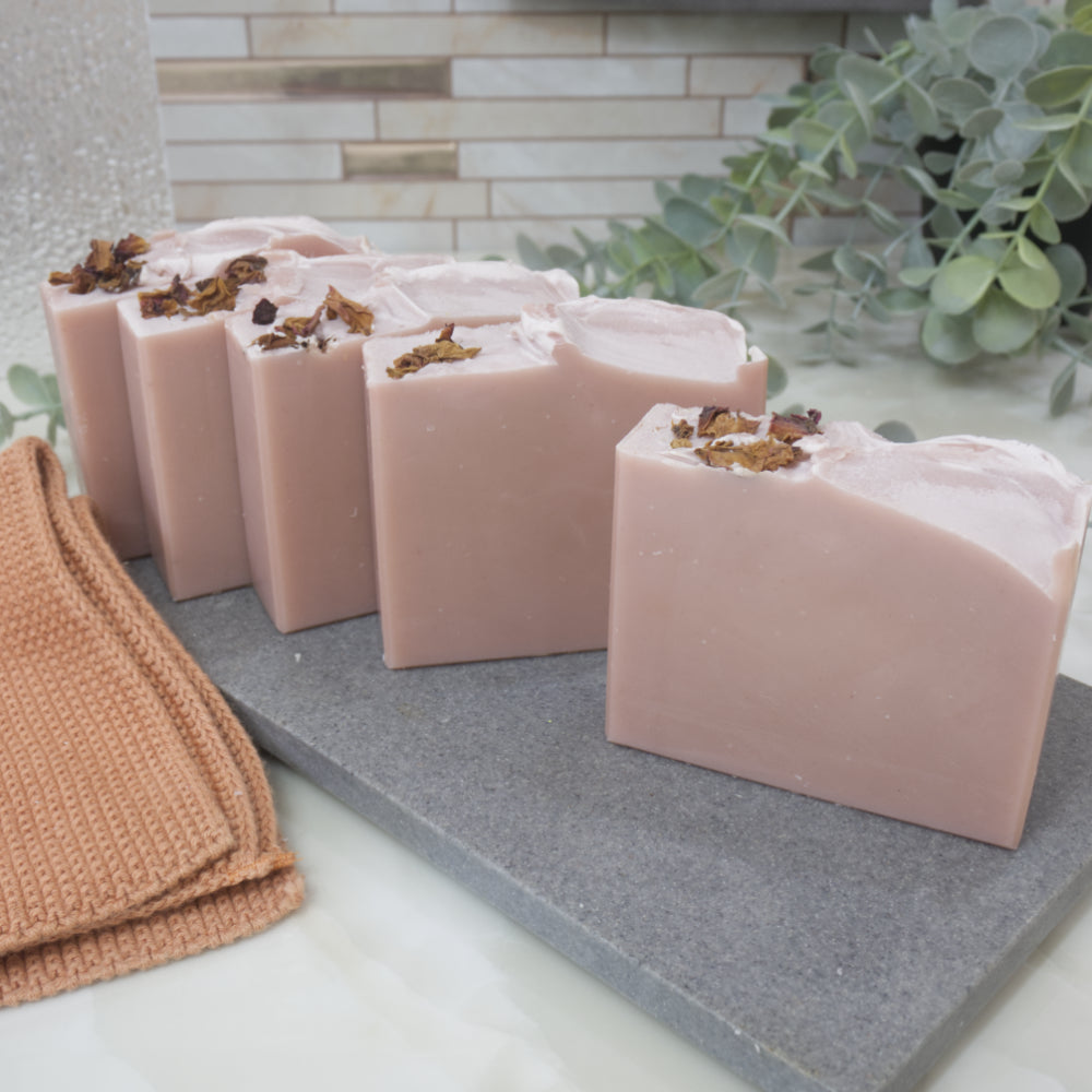 A selection of dusky pink soaps with rose petals on top sit on a grey stone surface against a tile background with greenery