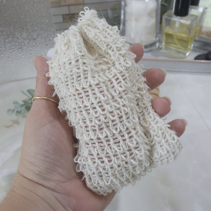 A Hand is holding a rough looking cotton bag against a blurred backdrop of a bathroom counter.   The bag is cream with a woven cotton like appearance with little loops on the bag