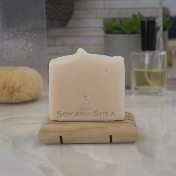 A cream coloured bar of Tropical Mango Clay-tanicals Soap sits on a wooden soap dish on a bathroom counter top with blurred background of bathroom accessories