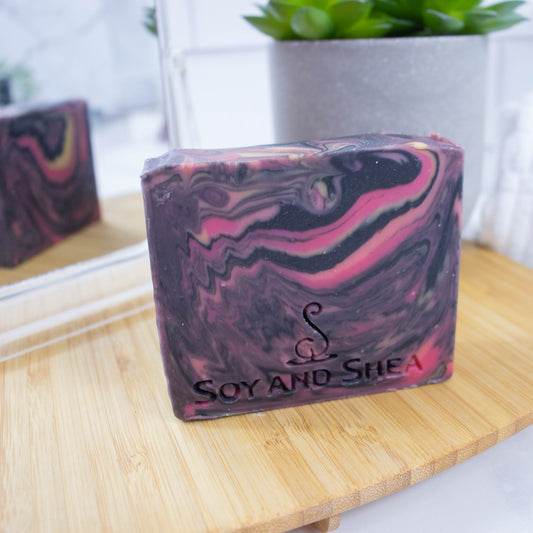 A bar of soap with a galaxy like swirl in black, pink and yellow stands on a wooden surface with a mirror reflecting the back of the soap showing a variation in the swirl design.