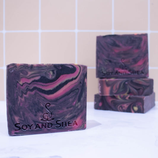 Marbled purple and pink soap bars with 'Soy and Shea' branding on a tiled bathroom background.