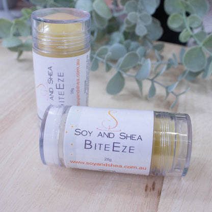 Two tubes of Soy and Shea's BiteEze Balm, a natural remedy for insect bites infused with lavender essential oil, sit on a wooden surface with a green plant in the background. The label on the nearest tube reads "BiteEze Balm 28g" and includes the website "soyandshea.com.au".