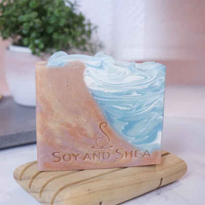 A soap bar sits on wooden soap dish in front of a potted plant with blurred background.  The soap features swirling patterns of colour with the left being in shade of brown and gold to look like sand and the right looking like water in shades of blue and white.