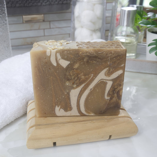 A rectangular bar of soap sits on a small wooden soap dish. The soap is a warm gold color with swirls of cream and dark gold. The background shows a softly blurred bathroom setting with a towel and a bottle.