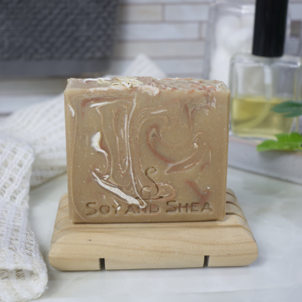 A bar of soap stands on a wooden soap rack next to a cotton cloth and bottle of perfume.  The soap has a honey coloured base colour with swirls of white and bronze throughout.   The top of the soap has pieces of rolled oats
