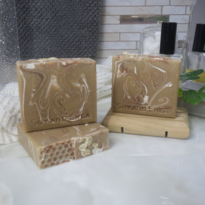 A selection of soaps are displayed on a bathroom countertop along with a towel, perfume bottle and mirror.  To the front right, a soap lays flat on the bench and with another standing upon it.  To the back right, a soap stands on a wooden soap rack.  The soaps have a golden honey base colour with various swirls of white and bronze throughout.  The top of the soap features a honeycomb effect on one half and some rolled oats on the other.