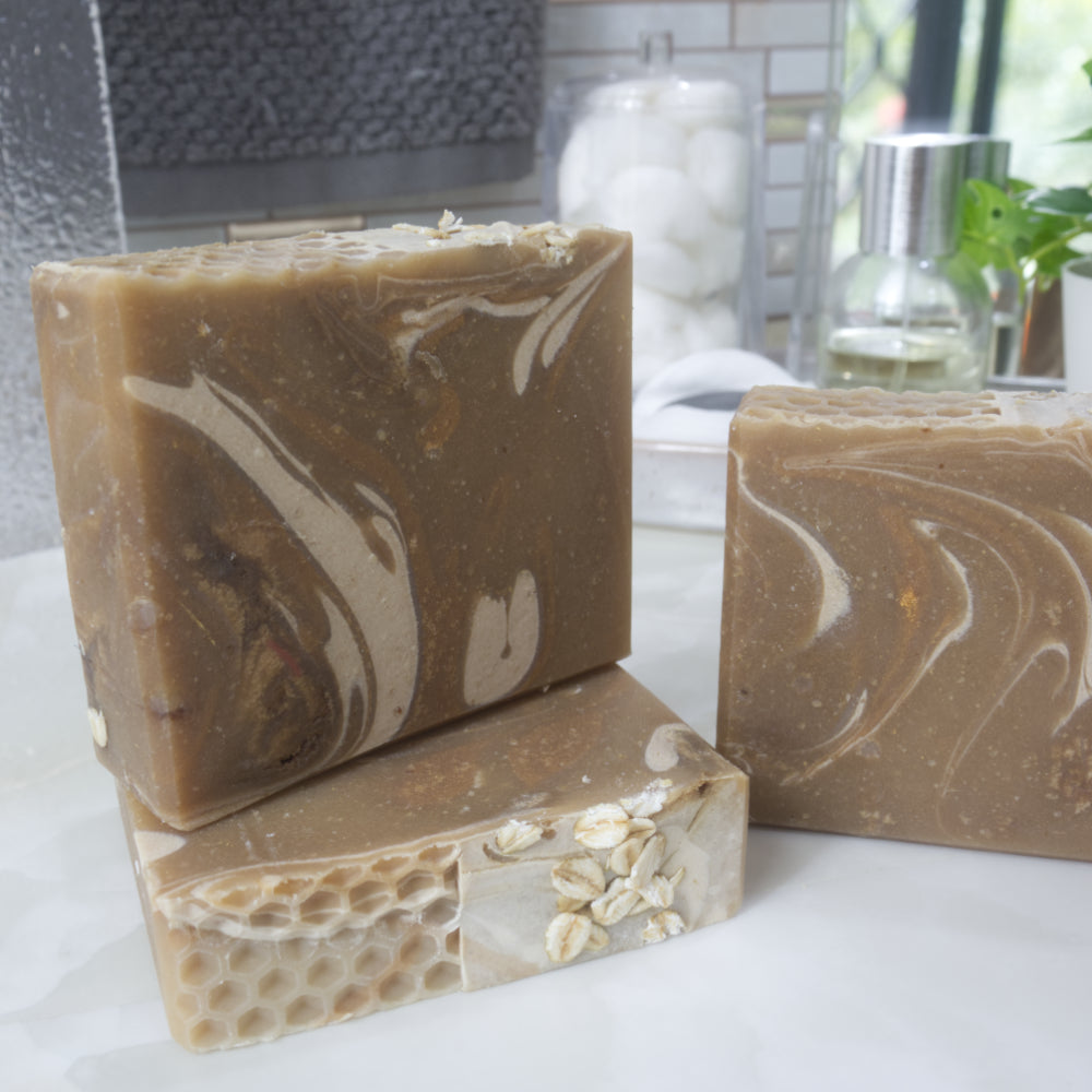 A collection of soaps are presented on a bathroom counter with blurred background.   One soap lays flat on the bench showing the soap top with a honeycomb pattern and oat pieces.  On top stands another soap and to the right is a glimpse of a third soap.  All soaps are a warm gold colour with swirls of Cream and rich gold