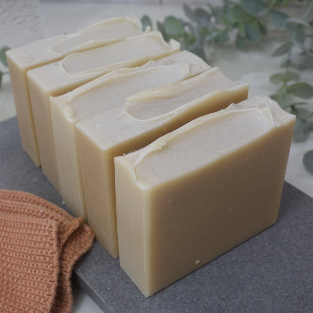 Five bars of soap sit in a row on a gray surface with a brown towel and green leaves in the background.  The soaps have a natural cream colour and some texture across the top