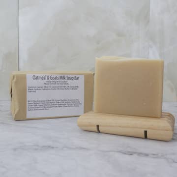 A dark cream soap bar sits on a wooden stand with a brown paper package behind labeled 'Oatmeal and Goats Milk Soap Bar' on a marble background