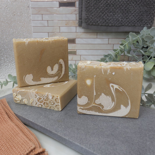 Three bars of brown soap with white drops of colour on a gray surface, with a tiled wall and plant in the background.  One bar of soap lays flat on the surface showing a top that has pieces of oat on one side adn a honeycomb pattern on the other