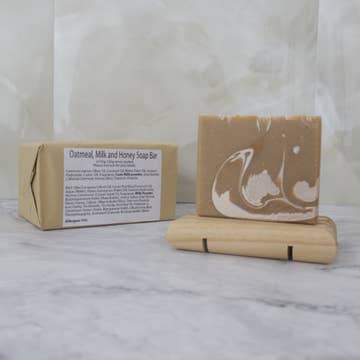 A tan soap bar with white drops sits on a wooden stand with a brown paper package behind labeled 'Oatmeal, mil and Honey Soap Bar' on a marble background
