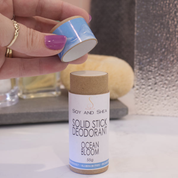 A cardboard tube labelled SOLID STICK DEODORANT stands upright on a bathroom counter top with a blurred background.  A hand hovers above the tube holding the lid in the finger tips revealing  a cream balm inside
