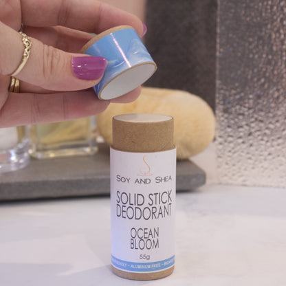 A cardboard tube labelled SOLID STICK DEODORANT stands upright on a bathroom counter top with a blurred background.  A hand hovers above the tube holding the lid in the finger tips revealing  a cream balm inside