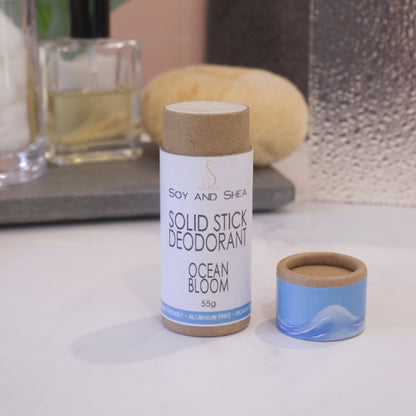 A cardboard tube labelled SOLID STICK DEODORANT stands upright on a bathroom counter top with a blurred background.  The lid of the tube sits to the left revealing the cream balm in the tube