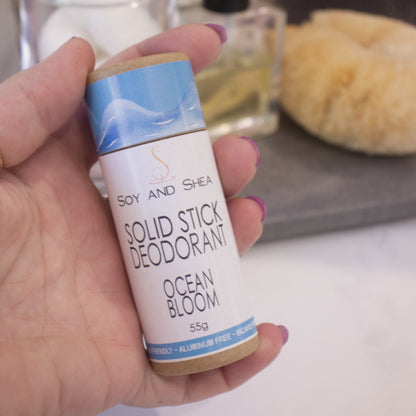 A hand holds a cardboard tube labelled as SOLID STICK DEODORANT against a blurred bathroom background. 