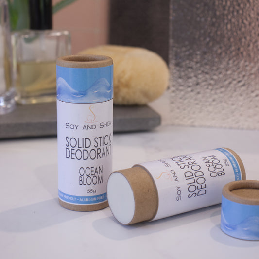 Two cardboard tubes labelled with SOLID STICK DEODORANT rest on a bathroom counter top with a blurred background.  One tube stands upright on the bench while another lays flat with the lid removed to reveal an off-white balm inside.  The label on the lid has a image of a pale blue wave against a bright blue background