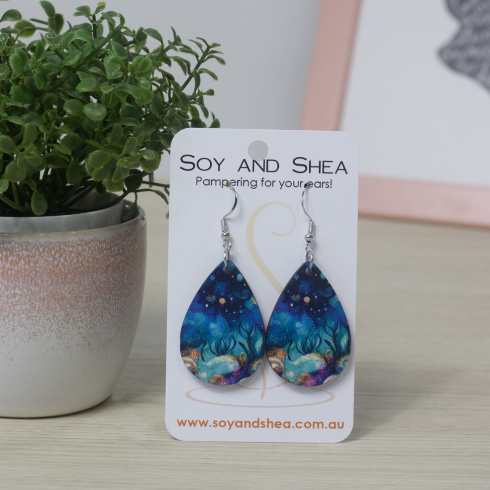Teardrop shaped earrings with a night underwater scene displayed on a Soy and Shea card with a plant in the background