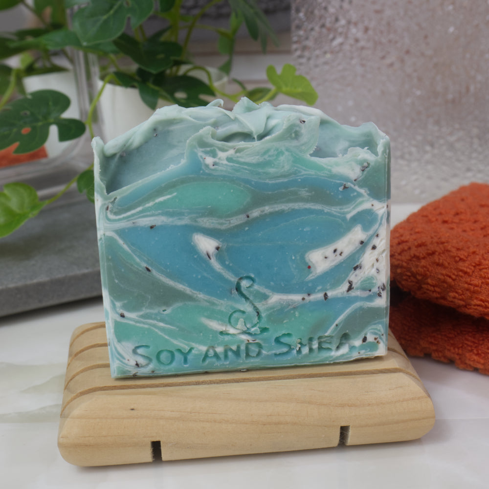 Marbled green and blue soap on a wooden stand with 'Soy and Shea' branding, plant and towel in the background.