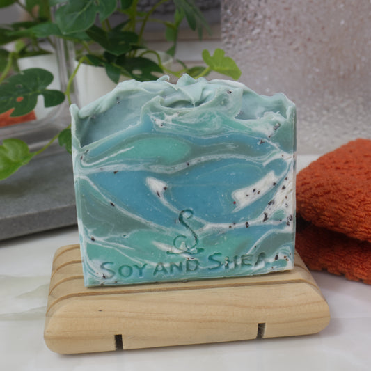 Marbled green and blue soap on a wooden stand with 'Soy and Shea' branding, plant and towel in the background.