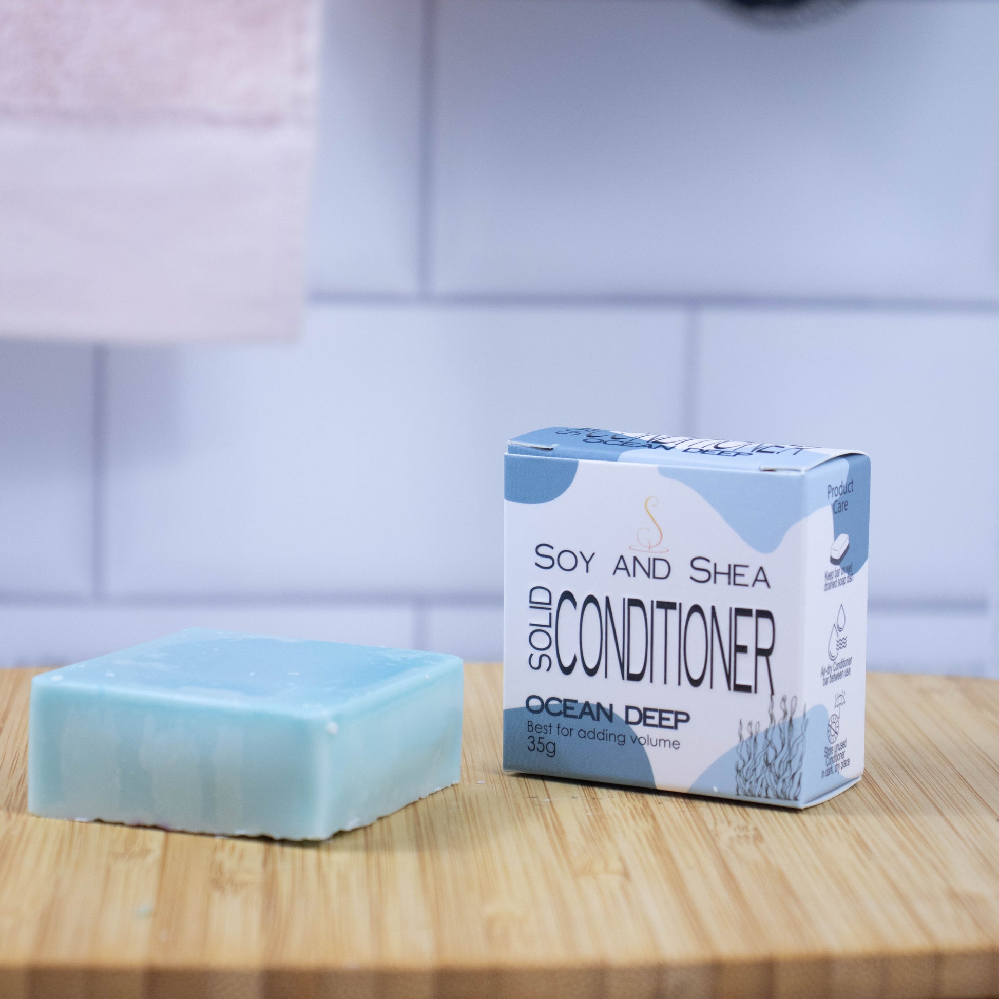 On a wooden tray sits a blue-green bar of conditioner next to a white box with a blue abstract pattern reading SOLID CONDITIONER against a blurred tiled background.