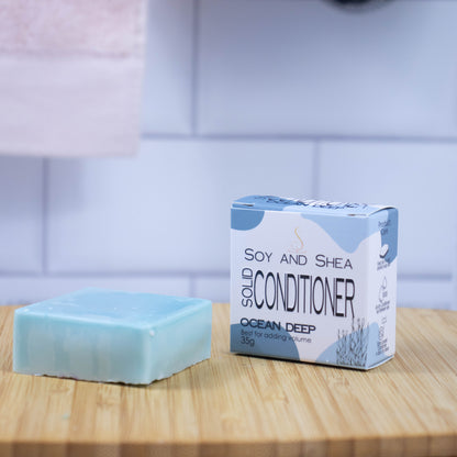 On a wooden tray sits a blue-green bar of conditioner next to a white box with a blue abstract pattern reading SOLID CONDITIONER against a blurred tiled background.