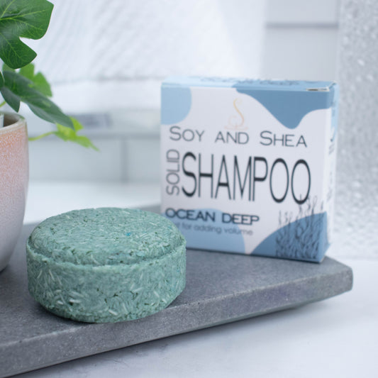 A white box with blue abstract pattern and the text SOLID SHAMPOO OCEAN DEEP stands upright on a grey stone tray with a blurred bathroom background.  Next to the box sits a round bar of solid shampoo.  The bar is ocean green with small white lines throughout.