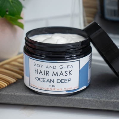 A black jar with the lid resting on its side revealing a white cream inside, sits on a dark grey tile surface against a blurred background of a bathroom counter top. The jar features a white label with blue abstract design with text HAIR MASK OCEAN DEEP.