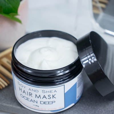 An overhead shot of a black jar with the lid resting on its side revealing a white cream inside, sits on a dark grey tile surface against a blurred background of a bathroom counter top. The jar features a white label with blue abstract design with text HAIR MASK OCEAN DEEP.