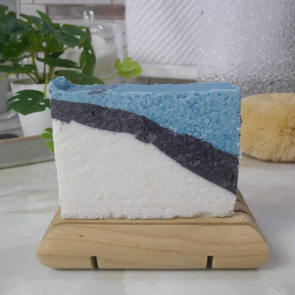 A soap bar stands on a wooden soap dish on a bathroom counter surounded by a plant, body brush and hanging towel.   The soap has a diagonal pattern that starts in the lower left as a large white area, followed by a narrow layer of black and finally a bright blue.